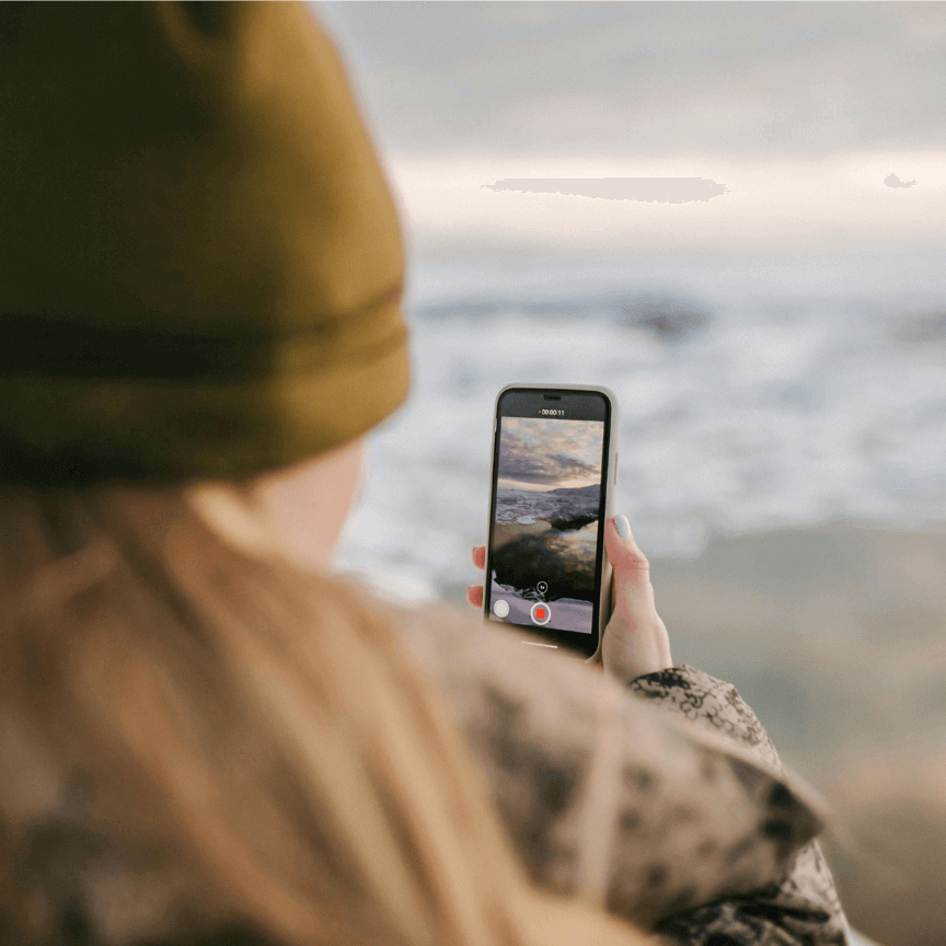 man in beanie hat stood facing the sea with phone in hand, filming